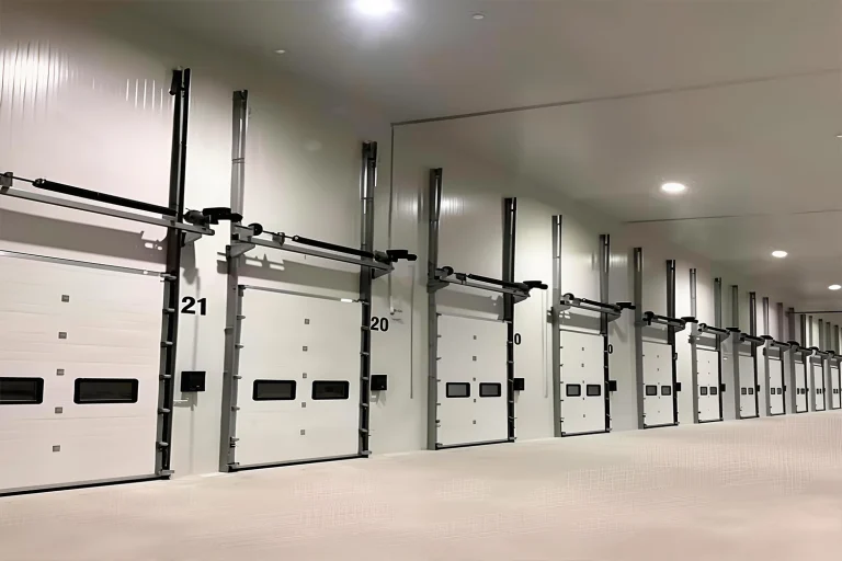 A long row of heavy-duty white vertical opening garage doors with vision panels inside a modern logistics hub, manufactured by ABX to maximize ceiling space for commercial projects.