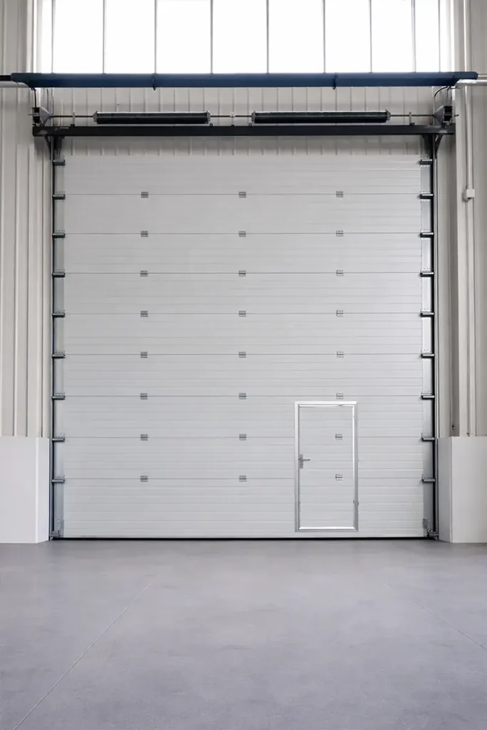 Interior view of an insulated industrial sectional door with a wicket door, showing robust vertical tracks and torsion spring systems.