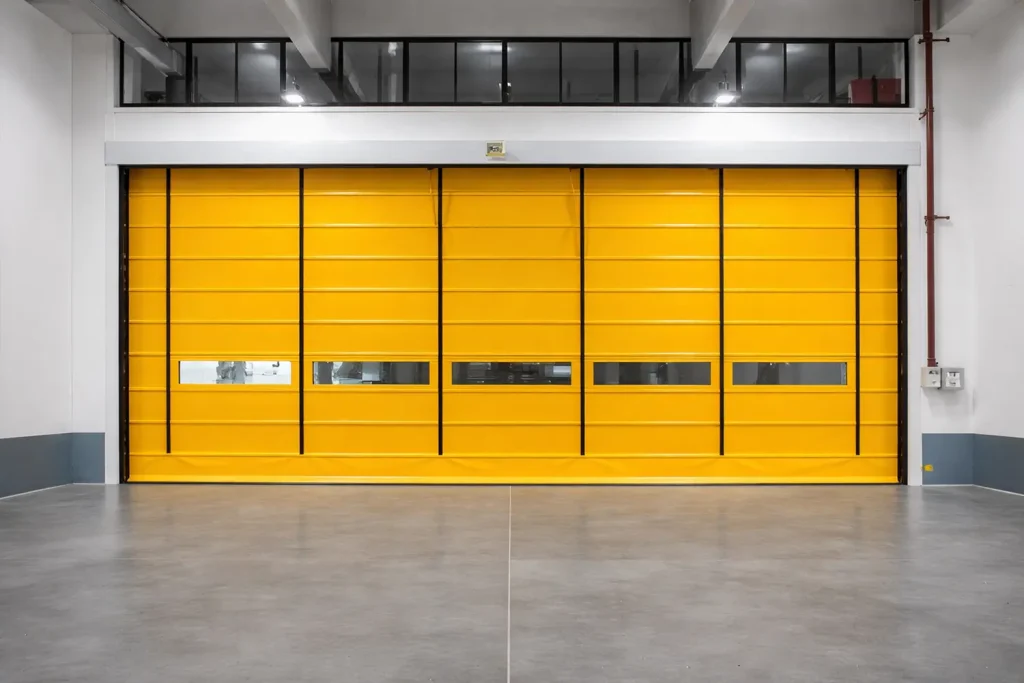 Wide yellow high-speed stacking door featuring prominent vertical lifting straps and segmented vision windows, used for environmental separation within a heavy manufacturing plant.